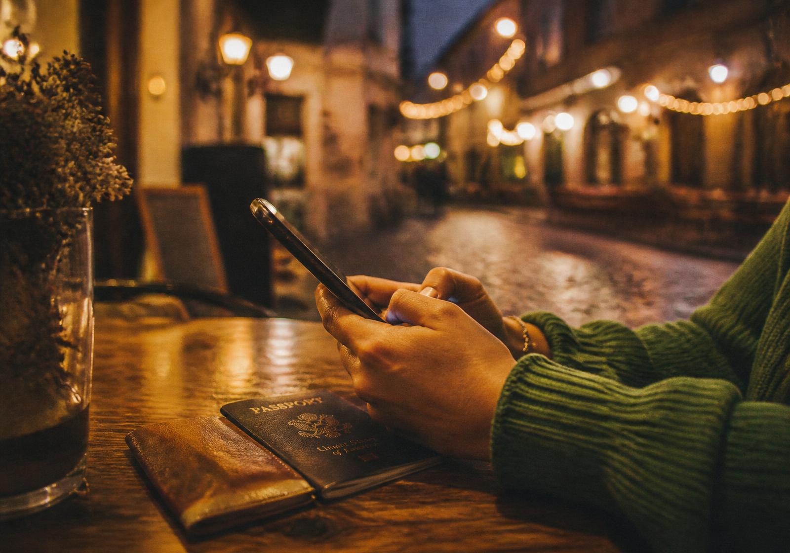Hände einer Frau halten ein Handy auf einem Cafe-Tisch, Reisepass daneben, warme Abendlichter im Hintergrund